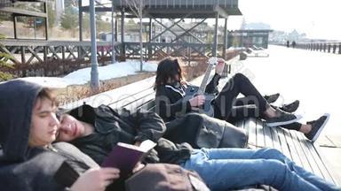 年轻的朋友坐在长凳上不寒而栗-两<strong>个人</strong>在看书-一<strong>个人</strong>在调吉他，他的女朋友在看他